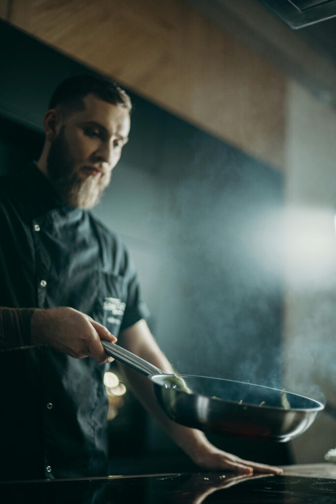 A focused chef prepares a meal using a frying pan in an indoor kitchen setting.