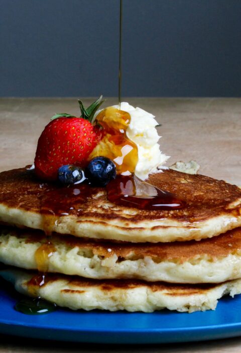 Stack of fluffy pancakes topped with syrup, strawberries, blueberries, and cream on a blue plate.