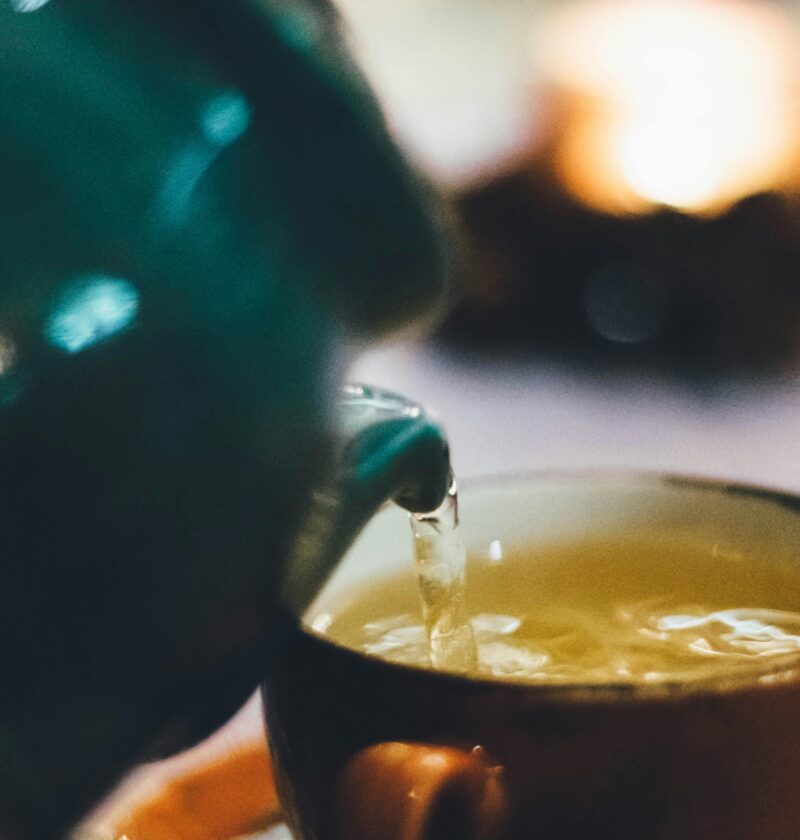 Warm close-up image of tea being poured into a teacup with blurred background ambiance.