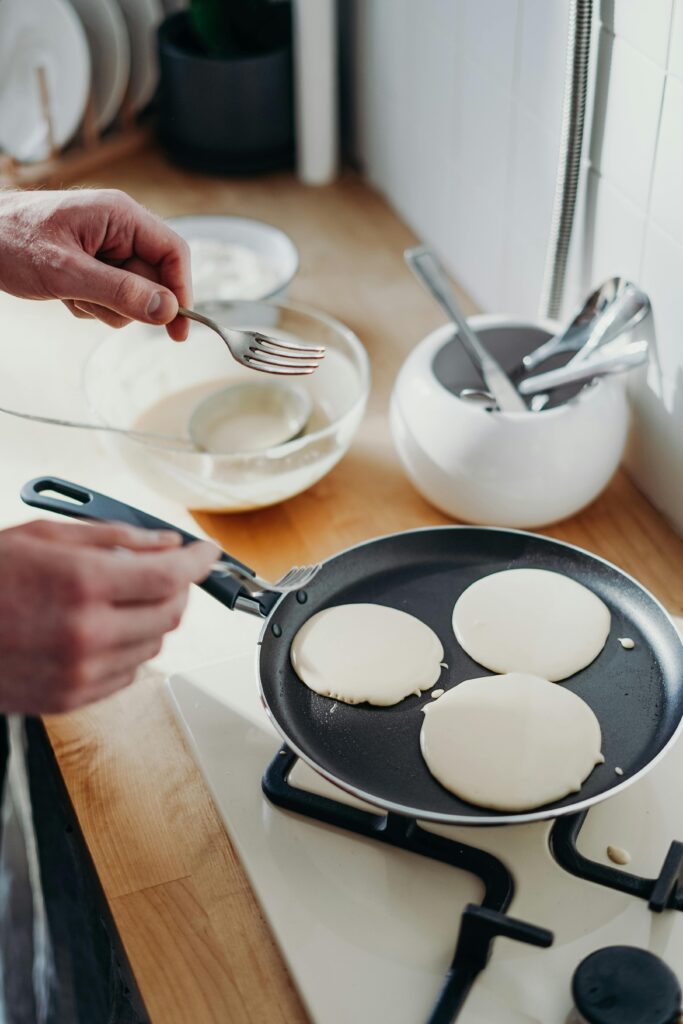 Close-up of pancakes cooking in a pan on a gas stove with utensils nearby.