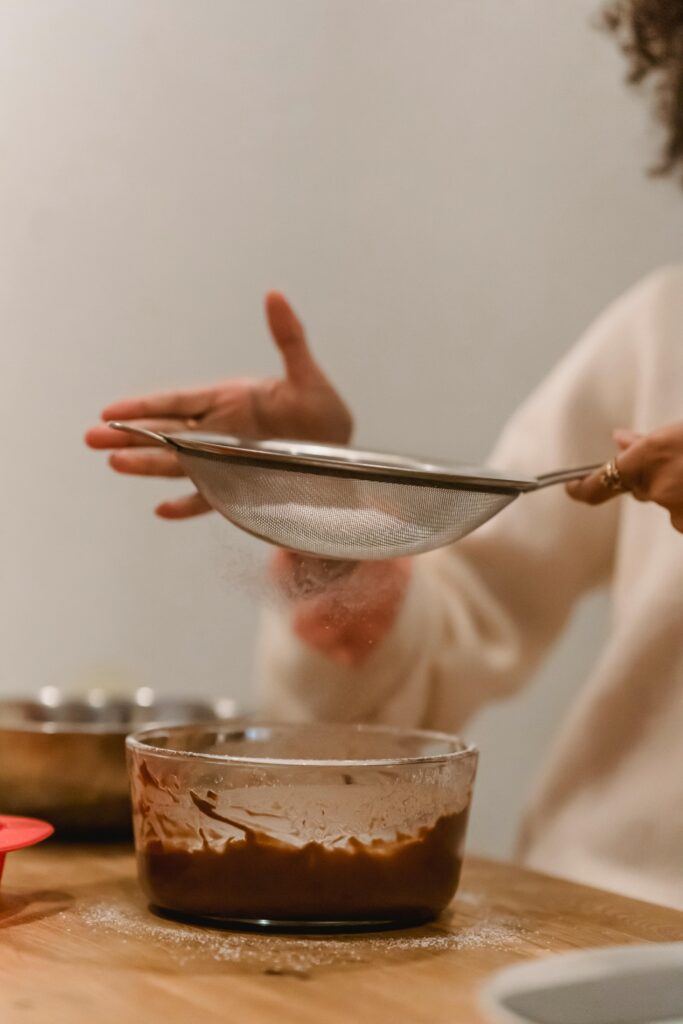 A woman sifts flour into a bowl of chocolate cake batter in a home kitchen.