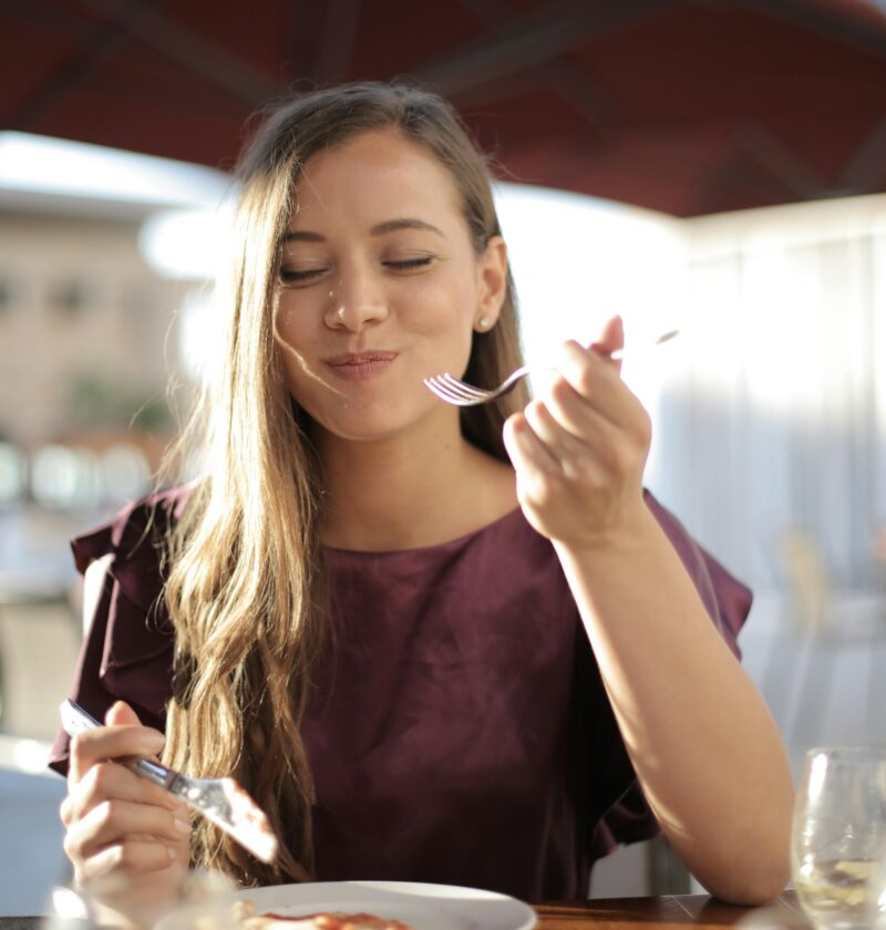A woman savoring her meal with a smile, dining outdoors in a pleasant setting.