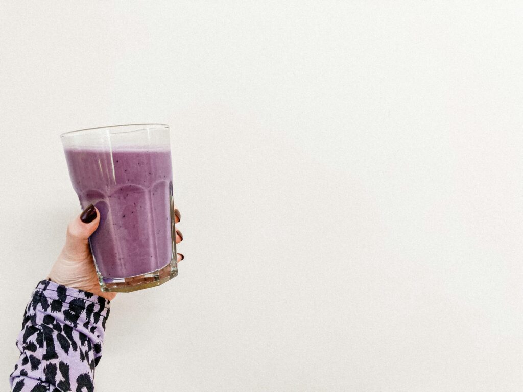 pexels photo 6435266 6435266 A person's hand holding a healthy purple smoothie in a glass with a white background. Perfect for health and wellness themes.